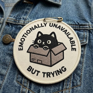 May include: Embroidered patch with a black cat peeking out of a cardboard box. The patch is circular with the words "Emotionally Unavailable" and "But Trying" in black text. The patch is attached to a denim jacket with a safety pin.