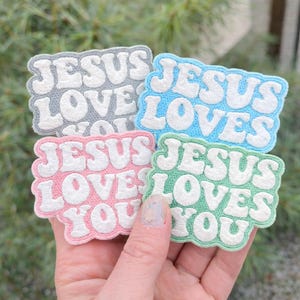 May include: Four embroidered patches in gray, blue, pink, and green, each with the words "JESUS LOVES YOU" in white, puffy letters. The patches have a scalloped edge and are held in a hand.
