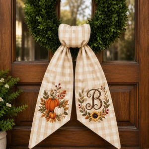 May include: A beige and white gingham fabric bow hanging from a green wreath. The left tail features an embroidered arrangement of orange and white pumpkins with autumn leaves. The right tail displays an embroidered letter B inside a floral wreath.