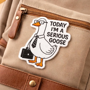 May include: A white patch featuring a cartoon goose wearing glasses, a tie, and carrying a briefcase. The patch has the text "TODAY I'M A SERIOUS GOOSE". The patch is attached to a tan backpack with brown leather accents.