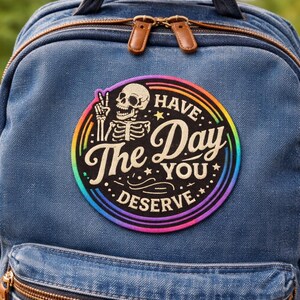 May include: Blue denim backpack with a brown zipper and leather zipper pulls. A circular patch features a skeleton giving a peace sign, surrounded by a rainbow and the words "Have The Day You Deserve."