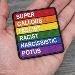 May include: A rectangular patch with a rainbow color scheme, featuring the words "SUPER," "CALLOUS," "FASCIST," "RACIST," "NARCISSISTIC," and "POTUS" in white block letters. The patch is held in a hand.