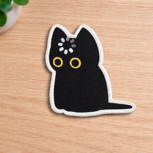 May include: Black cat patch with yellow eyes and a white border. The cat has a loading symbol on its head. The patch is on a light brown wooden surface.