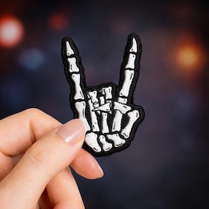 May include: A black and white embroidered patch in the shape of a skeleton hand making the rock and roll gesture. The patch has a black border and is held against a blurred background.