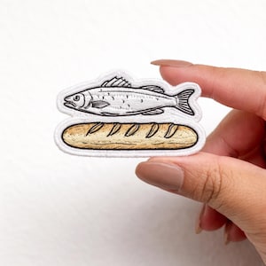 May include: Embroidered patch featuring a fish above a loaf of bread. The fish is white with black details, and the bread is tan with darker brown markings. The patch has a white border.