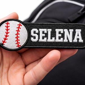 May include: A black patch with the name "SELENA" in white block letters. A baseball graphic is on the left side of the patch. The baseball is white with red stitching. The patch is held in a hand.