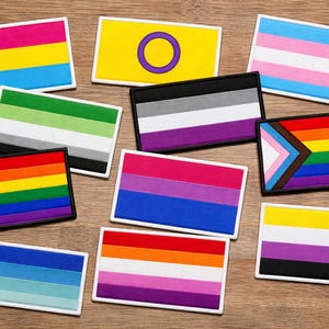 May include: A collection of rectangular patches featuring various LGBTQ+ pride flags. The patches display a range of colors, including rainbow, pink, blue, yellow, and purple, with white borders. The flags represent different identities and orientations.