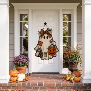 May include: A wooden ghost-shaped door hanger featuring a patchwork pattern of black, white, and orange plaid squares. The ghost is topped with a large patterned bow and holds a small orange jack-o'-lantern bucket filled with autumn foliage.