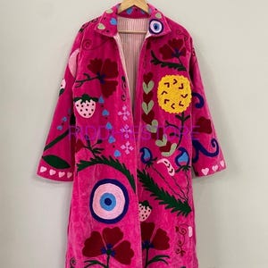 Könnte beinhalten: Ein langärmeliger, pinkfarbener Mantel mit lebendigen, mehrfarbigen Stickereien, darunter große rote Blumen, eine gelbe Sonne, Erdbeeren und blaue Auge-Motive. Das Kleidungsstück hat einen Kragen und hängt an einem Holzkleiderbügel.