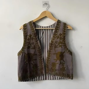 Könnte beinhalten: Eine Vintage-Weste mit floralem Design. Die Weste hat eine gedämpfte lila Farbe mit olivgrüner Stickerei und einem gestreiften Innenfutter. Sie hängt an einem Holzbügel vor einer weißen Wand.