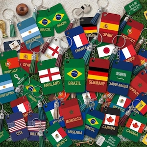 Peut inclure: Une collection de porte-clés rectangulaires arborant divers drapeaux nationaux et noms de pays, dont le Brésil, l'Argentine, l'Angleterre, l'Allemagne, l'Espagne et le Canada. Chaque porte-clés comprend un anneau métallique et un pompon.