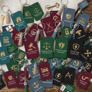 Peut inclure: Une collection de porte-clés rectangulaires verts, bordeaux et noirs, chacun arborant des icônes et du texte dorés représentant diverses professions comme comptables, électriciens et mécaniciens, accompagnés de pompons et d'anneaux métalliques.
