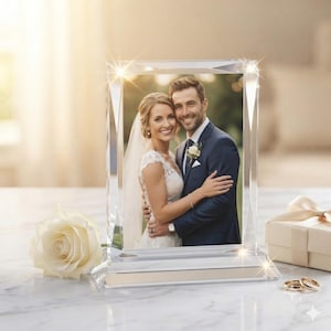Peut inclure: Un cadre photo rectangulaire en verre transparent aux bords biseautés contenant une photographie d'un couple en tenue de mariage. Le cadre est placé à côté d'une rose couleur crème, d'un petit coffret cadeau avec ruban et deux alliances en or.