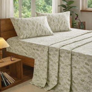 May include: A bed with a wooden frame is covered with a cream-colored sheet set featuring a green botanical pattern. Two matching pillowcases are visible. A wooden nightstand with a lamp and books sits beside the bed. The room has a window with white curtains.