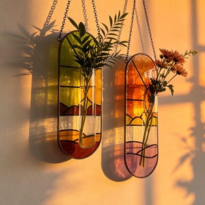 May include: Two oval stained glass wall planters with metal chains. The left planter features green, yellow, and red landscape panels holding green foliage. The right planter displays orange, yellow, and purple panels holding orange flowers.