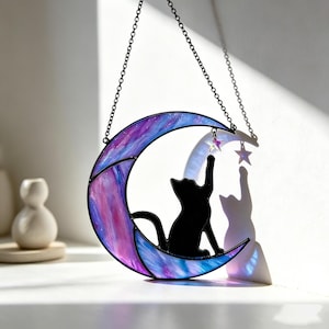 Puede incluir: Una luna creciente de vidriera con una silueta de gato negro que alcanza una estrella. La luna presenta una mezcla de tonos morados y azules. Una cadena suspende la pieza decorativa.
