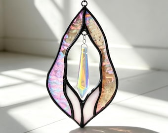 Puede incluir: Un atrapasoles de vidriera en forma de lágrima, con paneles de vidrio iridiscente rosa, dorado y blanco. Un cristal prismático transparente cuelga en el centro, reflejando los colores del arcoíris. La pieza está enmarcada con soldadura negra.