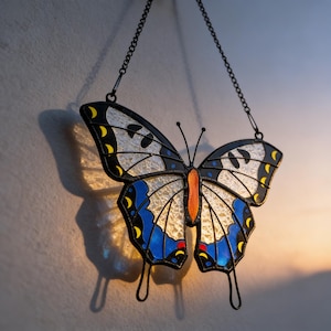 May include: A stained glass butterfly with black outlines, blue and white wings, and yellow accents. The butterfly is suspended by a black chain, casting a shadow on the wall. The butterfly's body is orange.