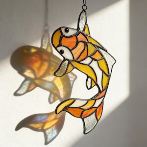Puede incluir: Atrapasoles de pez koi de vidriera con segmentos naranjas, amarillos y blancos, delineados en negro. El pez está suspendido por una cadena, proyectando una sombra colorida. Una pieza decorativa para el hogar.