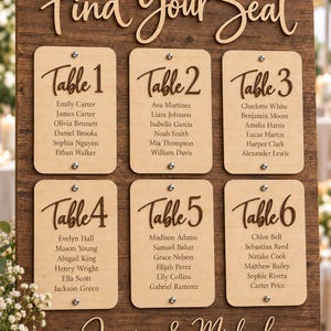 May include: A dark wood wedding seating chart sign with the text Find Your Seat and Jessica & Michael October 14, 2025. It features six light wood rectangular panels labeled Table 1 through Table 6, each listing guest names in dark brown script.