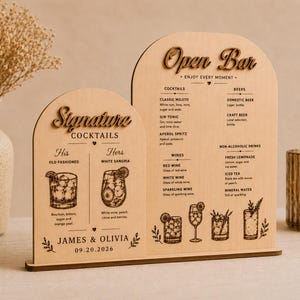 May include: Two arched light wood signs featuring laser-engraved text and illustrations. The smaller sign reads Signature Cocktails, His Old Fashioned, Hers White Sangria, James & Olivia, 09.20.2026. The larger sign reads Open Bar, Enjoy Every Moment.