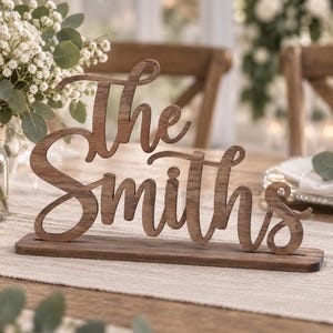May include: A dark brown wooden sign featuring the words The Smiths in a cursive script font. The sign is mounted on a rectangular wooden base and stands upright, displaying a natural wood grain texture throughout the lettering and the base.