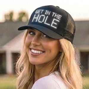 May include: A black trucker-style baseball cap featuring a mesh back and a solid front panel. The front of the cap displays the text GET IN THE HOLE in white, capitalized, sans-serif lettering. The cap has a curved brim and an adjustable strap.