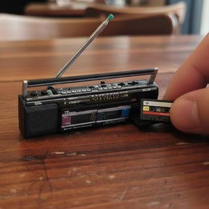 May include: Miniature black boombox with a silver antenna and a cassette tape. The boombox has a speaker, buttons, and a handle. The cassette tape is black, red, and yellow. The boombox is on a wooden surface.