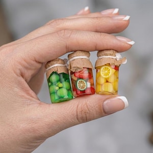 Puede incluir: Tres tarros de vidrio en miniatura llenos de fruta colorida. Un tarro contiene esferas verdes, otro tiene fresas rojas y una rodaja de lima, y el último contiene fruta amarilla y una rodaja de limón. Cada tarro está sellado con una tapa de papel marrón.
