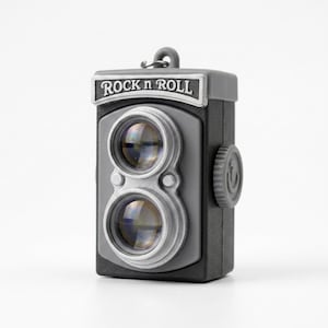 May include: A black and gray camera-shaped keychain featuring two prominent circular lenses and a silver-toned rectangular plate at the top with the text ROCK n ROLL embossed in black lettering. The device includes a side adjustment dial.