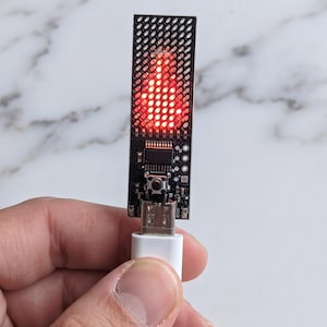 May include: A rectangular black circuit board device featuring a grid of small LED lights that are illuminated in red to form a flame-like pattern. The electronic component includes a central push button and a silver USB connector at the base.