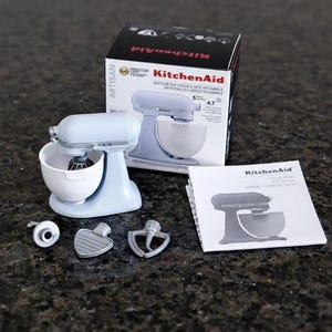 May include: A light blue KitchenAid Artisan stand mixer with a white bowl and attachments. The mixer is displayed with its box and a manual on a dark speckled surface. The box features the KitchenAid logo and product details.