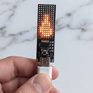以下が含まれることがあります： 小さな穴のグリッドと炎の形に光るオレンジ色のLEDディスプレイを備えた、長方形の黒い回路基板デバイス。この電子部品には、中央の押しボタンと下部に銀色のUSBコネクタが付いています。