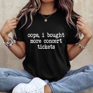 Könnte beinhalten: Ein schwarzes kurzärmeliges T-Shirt mit dem weißen Schriftzug "oops, i bought more concert tickets" in einer einfachen serifenlosen Schrift auf der Brust. Das T-Shirt hat einen Rundhalsausschnitt und wird mit blauen Jeans und weißen Turnschuhen kombiniert.