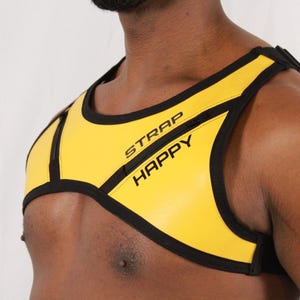 May include: Yellow harness with black trim and the text "STRAP HAPPY" in black. The harness is designed to be worn around the chest and shoulders. The background is a plain white.