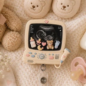 May include: A badge reel featuring a cream-colored monitor displaying an ultrasound image with miniature baby-themed charms, attached to a clear plastic card holder with a white insert that reads tiny MIRACLE in pink and brown text.