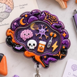 Op de afbeelding: Een hersenvormige badgehouder in paars en oranje met Halloween-bedels zoals een schedel, een neuron, een reflexhamer en een pillenflesje met pompoenafbeelding, bevestigd aan een doorzichtige plastic clip met een uittrekbaar koord.
