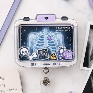 May include: A rectangular badge reel featuring a clear window with a miniature X-ray of a human rib cage, decorated with small floating charms including a skull, bone, radiation symbol, and stethoscope, labeled X-RAY VIEWER RADIOLOGY TECH.