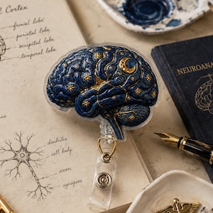 Puede incluir: Un portacredenciales con una ilustración de un cerebro azul oscuro decorado con estrellas doradas y una luna creciente. El diseño del cerebro se superpone a una página de cuaderno con diagramas anatómicos de la corteza cerebral, el sistema límbico y una neurona.
