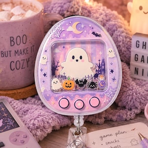 May include: A purple oval-shaped badge reel featuring a shaker-style display with a central ghost graphic, surrounded by small floating icons of ghosts, a pumpkin, a bat, a spider, and a potion bottle, with three circular buttons at the bottom.