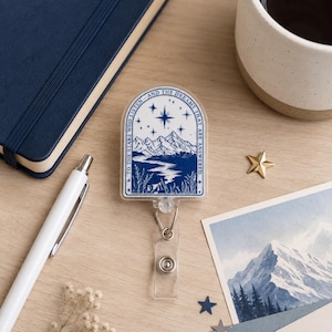 May include: A blue and white badge reel featuring a mountain landscape and the text "TO THE STARS WHO LISTEN AND THE DREAMS THAT ARE ANSWERED". It rests on a wooden surface with a pen, notebook, and a gold star.