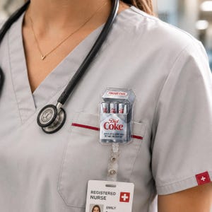 May include: A clear, plastic badge reel featuring a miniature Diet Coke cigarette pack design. The pack reads "Diet Coke" with the text "FRIDGE CIGS" and "JUST A LITTLE SOMETHING TO TAKE THE EDGE OFF". A stethoscope and a nurse's badge are also visible.