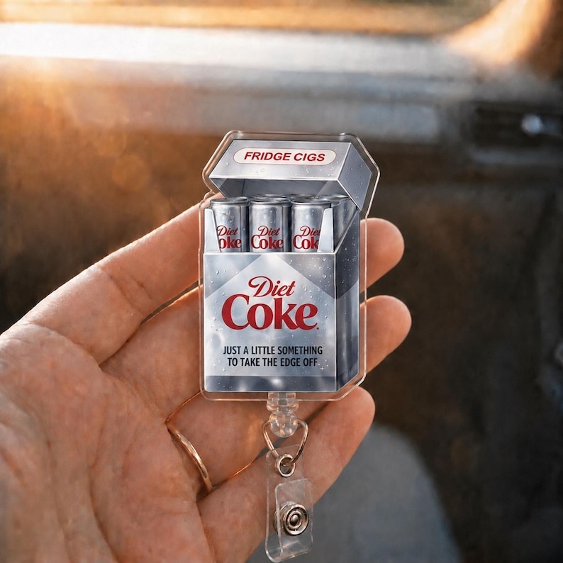 May include: A clear acrylic badge reel featuring a Diet Coke-themed design. The design mimics a cigarette pack labeled "FRIDGE CIGS" with Diet Coke cans inside. The text "Diet Coke" and "JUST A LITTLE SOMETHING TO TAKE THE EDGE OFF" are also visible.