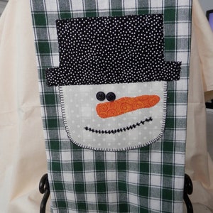May include: A green and white checkered kitchen towel with a snowman face appliqué. The snowman has a black top hat, orange nose, and black eyes.