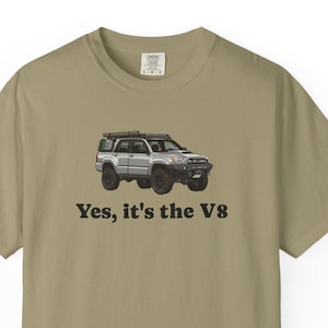 May include: A khaki-colored short-sleeve t-shirt featuring a graphic of a silver off-road SUV with a roof rack. Below the vehicle, the text "Yes, it's the V8" is printed in a bold, black, serif font. The shirt has a crew neckline and a standard fit.