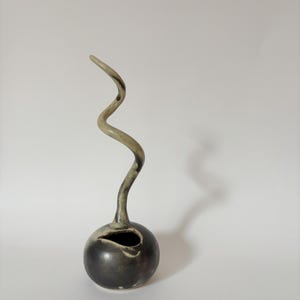 Puede incluir: Un jarrón de cerámica gris oscuro con una base bulbosa y un tallo largo y curvo de color claro. El jarrón tiene una pequeña abertura en el lateral de la base. El tallo se curva hacia arriba y hacia la izquierda.