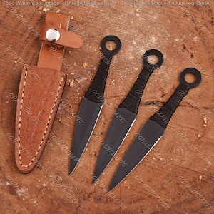 May include: Three black metal throwing knives with ring pommels and cord-wrapped handles, accompanied by a tan leather sheath featuring a silver-toned snap button closure. The knives have pointed blades and a uniform dark finish throughout.