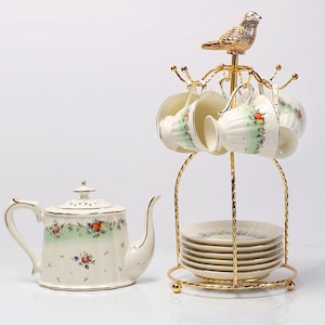 Op de afbeelding: Een crèmekleurige porseleinen theeset met bloemmotieven en gouden accenten. De set bevat een theepot, zes kopjes en schotels, gepresenteerd op een goudkleurige metalen standaard met een vogelbeeldje. De theepot heeft een gebogen handvat en tuit.