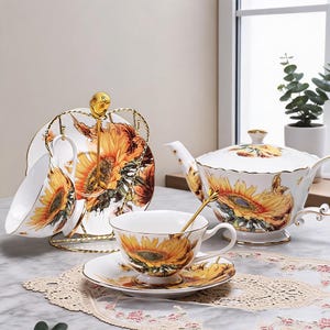 Puede incluir: Juego de té de porcelana blanca con diseño de girasoles. El juego incluye una tetera, tazas, platillos y un soporte. Los girasoles son amarillos, naranjas y marrones con detalles dorados.