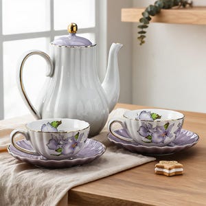 Puede incluir: Un juego de té de porcelana blanca que incluye una tetera con tapa color lavanda y borde dorado, acompañado de dos tazas y platillos a juego decorados con motivos florales morados. Una pequeña galleta en forma de estrella descansa al lado.